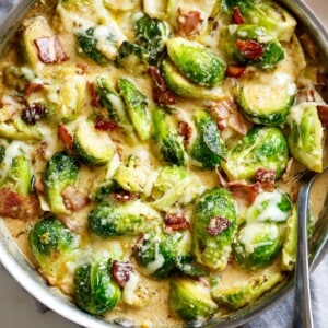 Creamy Garlic Parmesan Brussel Sprouts & Baconย in a silver frying pan with blocks of parmesan cheese in the background. Pan sits on a grey tea towel on a white marble background with a silver serving spoon in the pan. Your NEWย favoriteย way to eatย Brussels Spouts!
