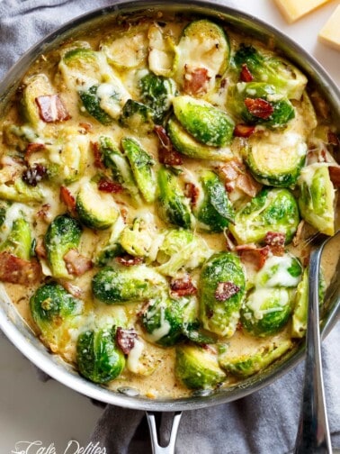 Creamy Garlic Parmesan Brussel Sprouts & Bacon in a silver frying pan with blocks of parmesan cheese in the background. Pan sits on a grey tea towel on a white marble background with a silver serving spoon in the pan. Your NEW favorite way to eat Brussels Spouts!