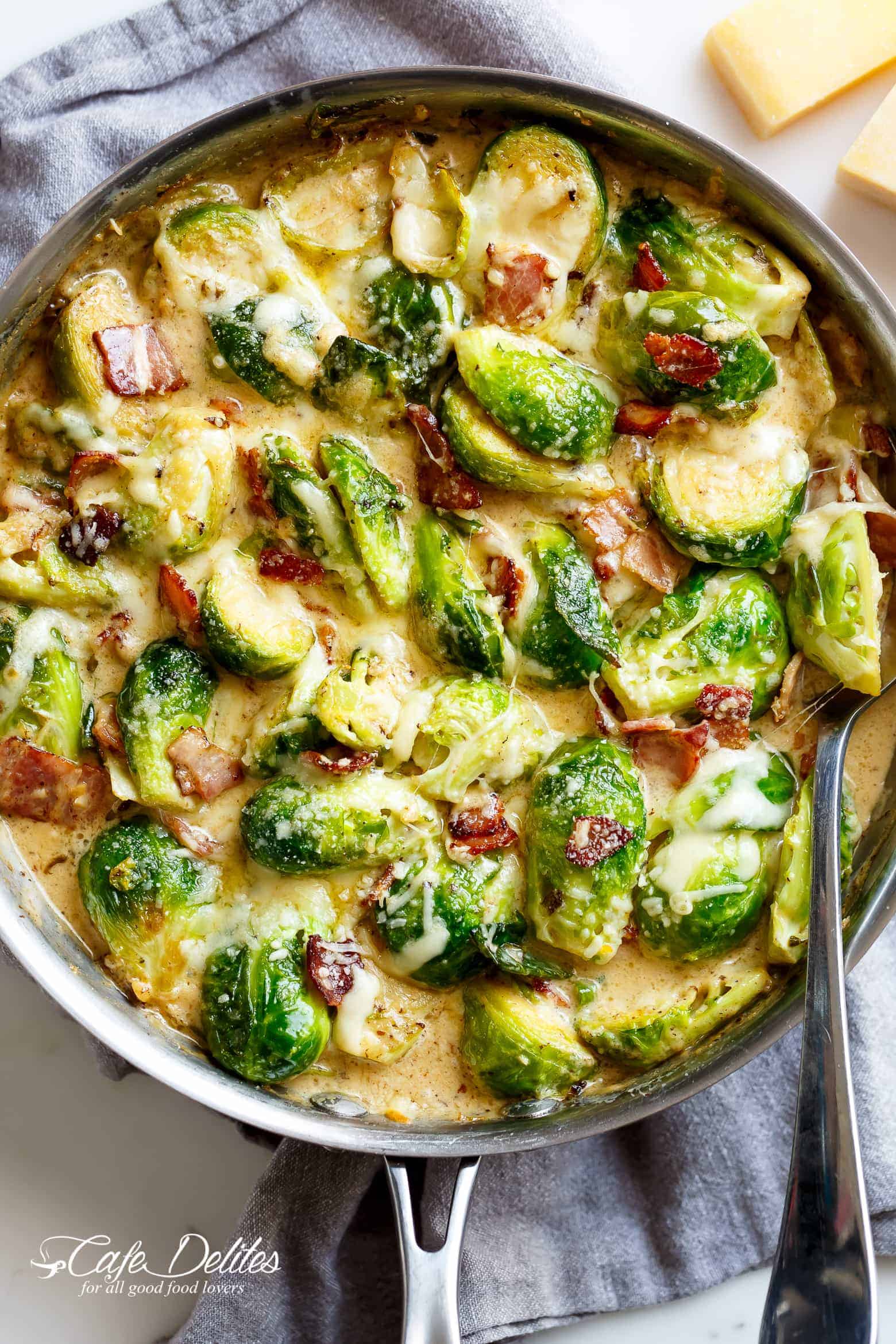 Creamy Garlic Parmesan Brussel Sprouts & Bacon in a silver frying pan with blocks of parmesan cheese in the background. Pan sits on a grey tea towel on a white marble background with a silver serving spoon in the pan. Your NEW favourite way to eat Brussels Spouts! 
