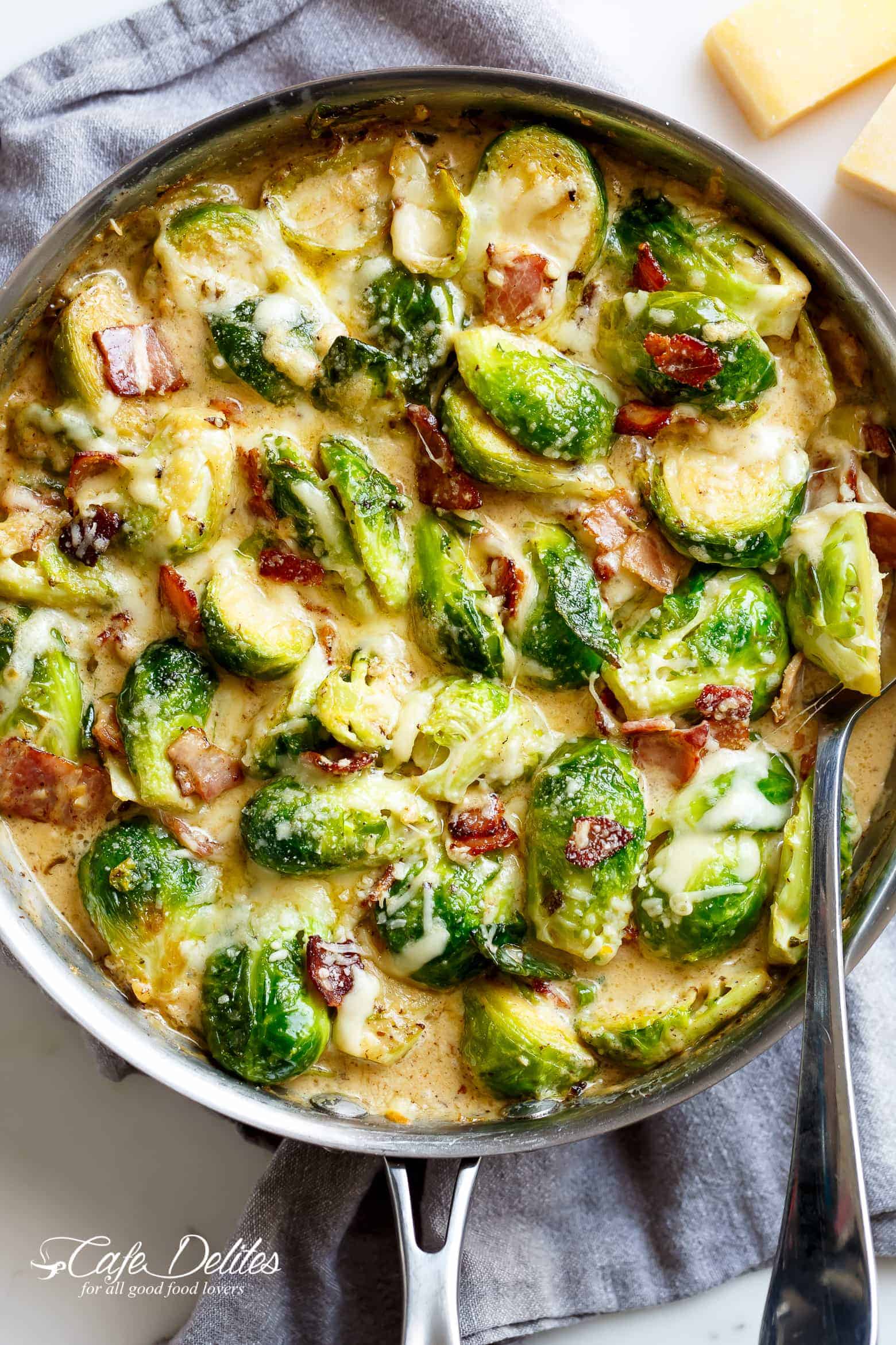 Creamy Garlic Parmesan Brussel Sprouts & Bacon in a silver frying pan with blocks of parmesan cheese in the background. Pan sits on a grey tea towel on a white marble background with a silver serving spoon in the pan. Your NEW favorite way to eat Brussels Spouts! 