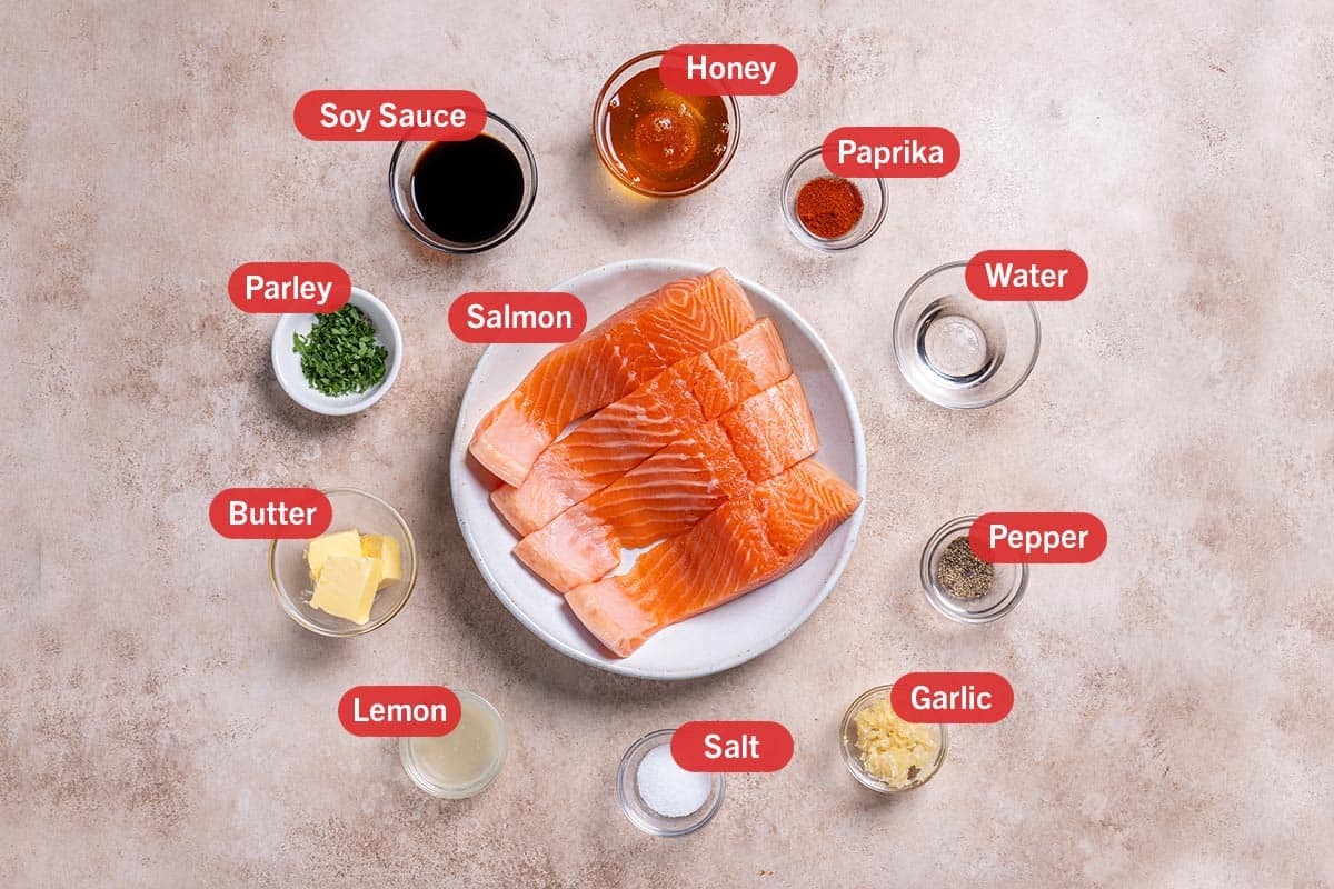 Image of the ingredients needed for the preparation of this recipe, specifically: salmon, soy sauce, honey, paprika, water, pepper, garlic, salt, lemon, butter, and parsely.