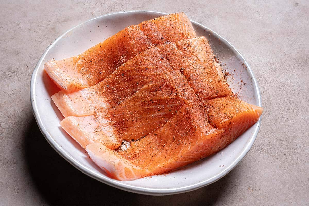 An image depicts four salmon fillets being seasoned in a white bowl while the oven is preheating.