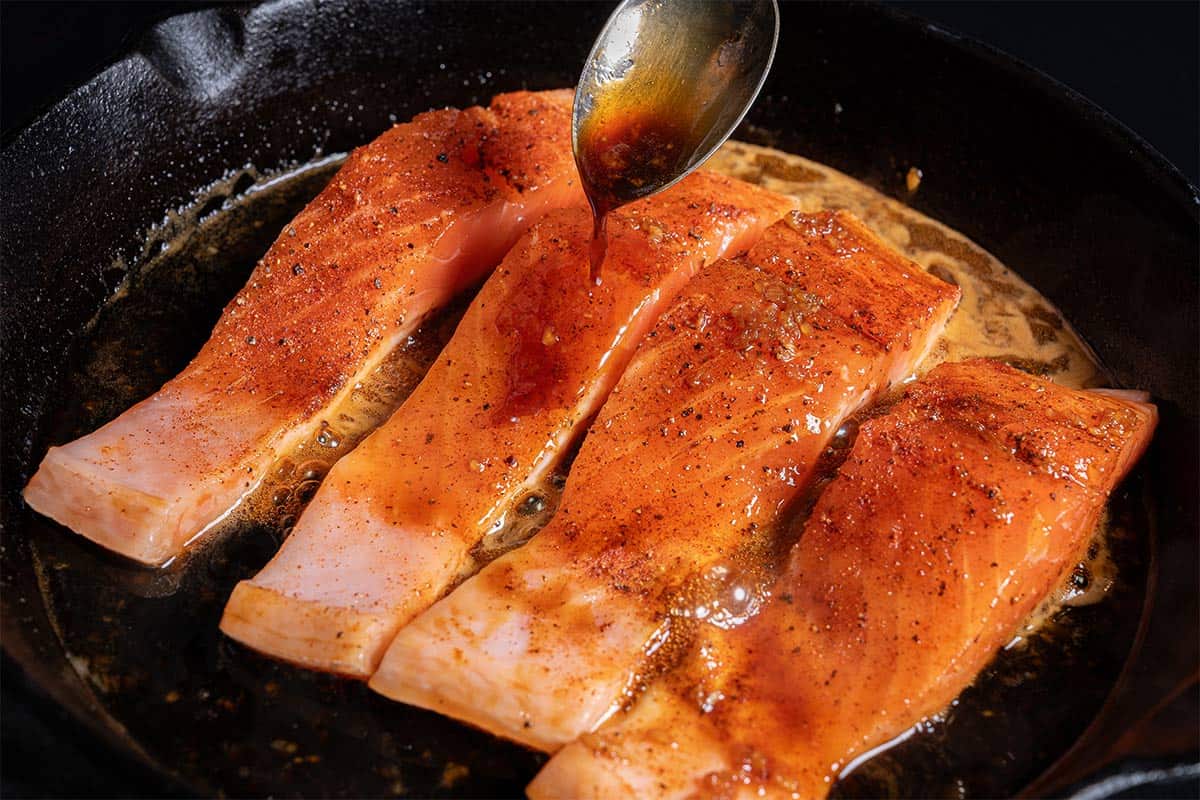 An image of salmon being cooked in a skillet with sauce. A spoonful of soy sauce is being added to the fillets as they cook.