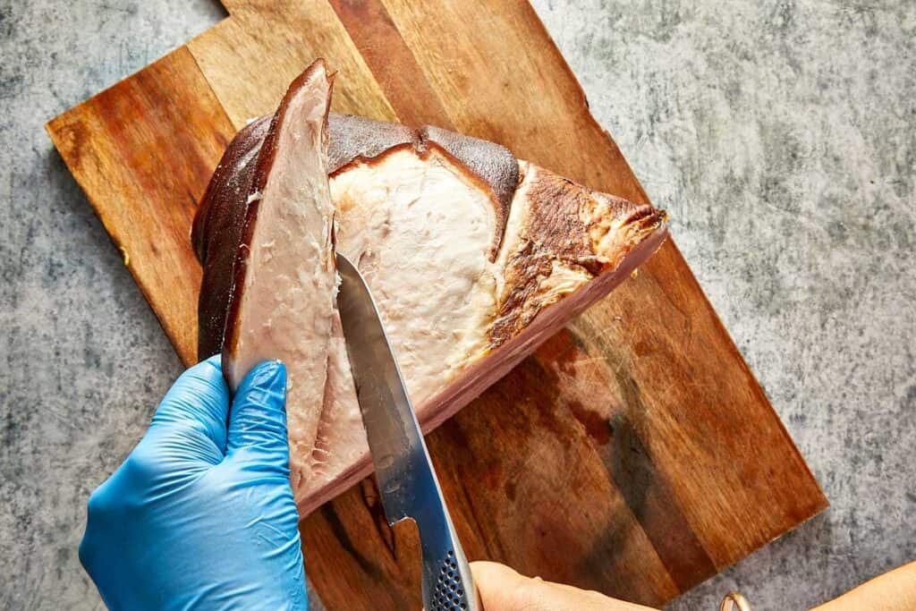 A top down view of a leg of ham on a wooden chopping board with a knife removing the rind off the leg ham exposing a white layer of fat - 5
