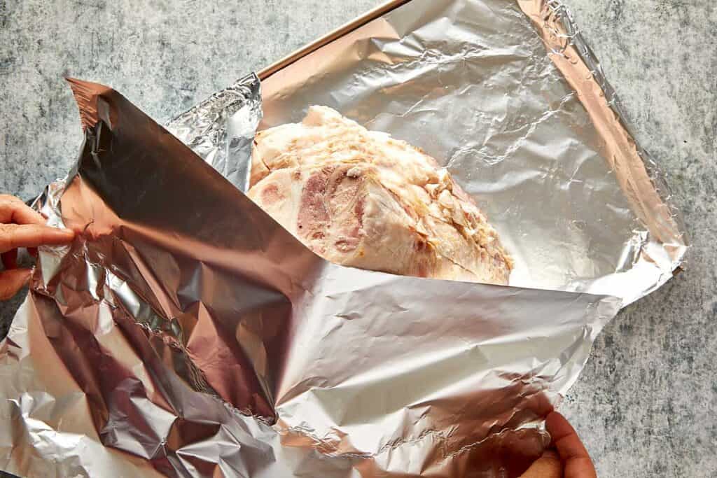 a top down view of a leg ham sitting on aluminum foil, the leg is sitting cut side down looking like a mountain from top down and exposing the fatty layer and foil covering the top of the ham - 12