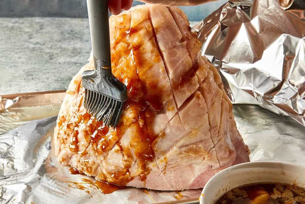 a side view of leg ham with a brush coating it with a glaze and a bowl of glaze on the side. - 15