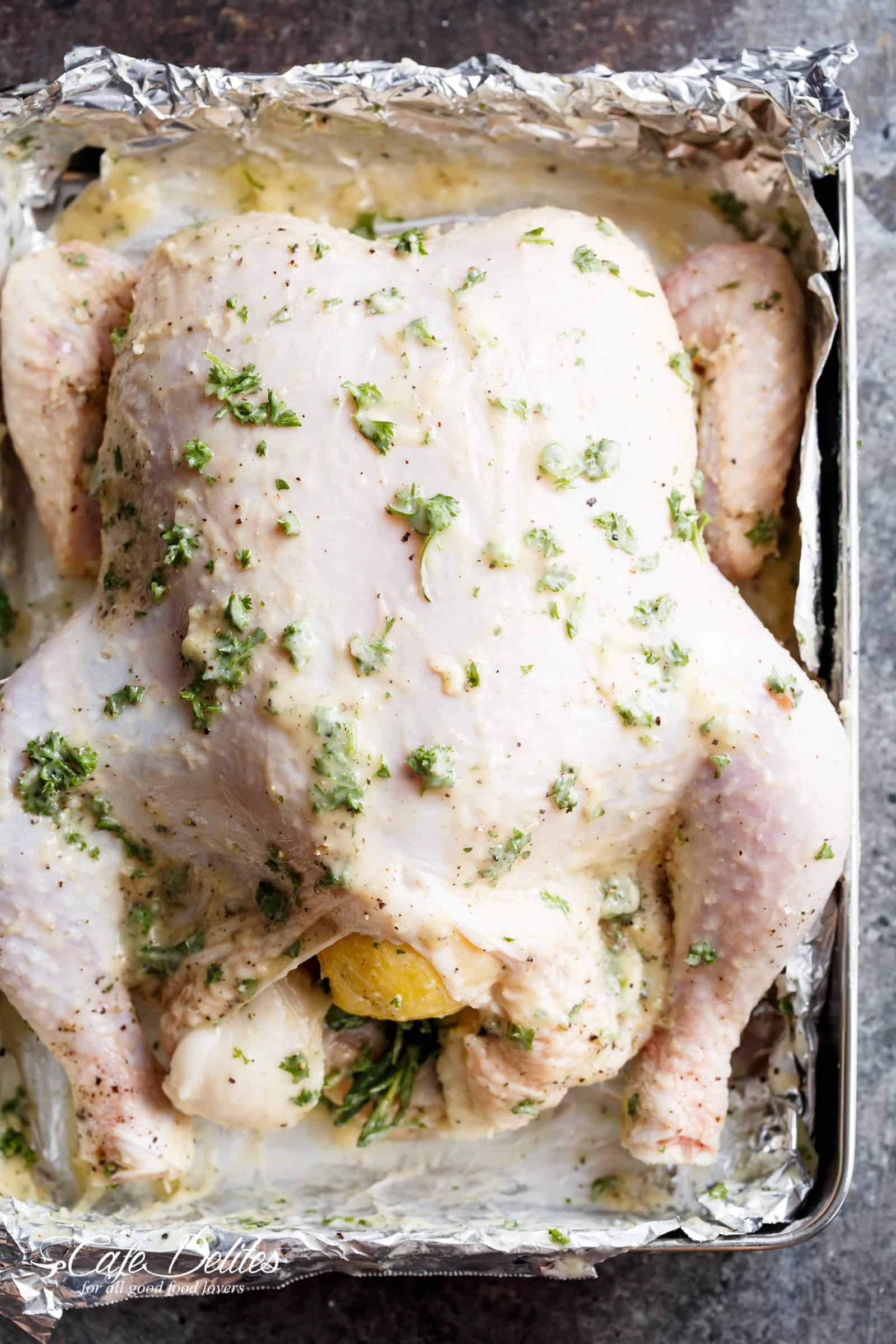 A whole, stuffed chicken slathered with garlic butter, rosemary, parsley, lemon, and a hint of white wine for an unbeatable flavour, laid in a tin foil lined pan ready for roasting.