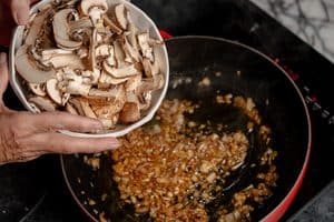A close-up view of a person's hands pouring a white bowl full of sliced raw brown mushrooms into a red skillet containing sizzling, browned chopped onions on a black electric stovetop.