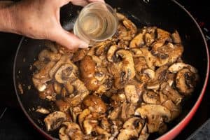 A close-up view of a person's hand pouring a small glass of clear liquid into a red skillet filled with steaming, sautéed sliced mushrooms and onions on a black stovetop.