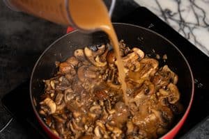 A close-up view of a brown liquid being poured from a clear pitcher into a red skillet filled with steaming, sautéed sliced mushrooms and onions on a black electric stovetop.