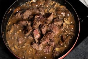 A close-up view of seared, partially cooked beef cubes piled in the center of a rich brown mushroom and onion gravy inside a red skillet on a black stovetop.
