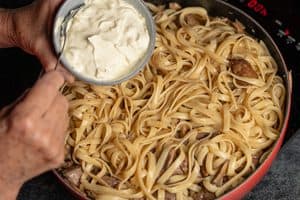 A close-up, top-down view of a person's hands holding a small bowl of thick white cream with a spoon, poised over a red skillet completely filled with cooked flat pasta resting on top of a beef and mushroom mixture on a black electric stovetop.