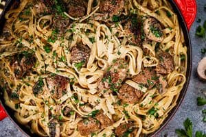 A close-up, top-down view of a finished beef and mushroom stroganoff inside a red skillet, featuring flat pasta, seared beef chunks, and sliced mushrooms coated in a creamy brown sauce and generously garnished with chopped fresh parsley and black pepper.