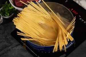 A close-up view of long strands of dry pasta being added into a blue pot of steaming, boiling water on a black electric stovetop with a red digital display, with fresh herbs and raw beef slightly blurred in the background.
