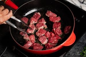 A close-up view of a person's hand using metal tongs to turn small cubes of raw and partially browned beef searing in oil inside a bright red skillet on a black electric stovetop.