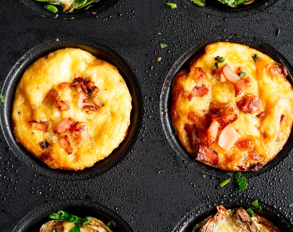 A muffin tray with three rows of different flavored breakfast muffins. One row contains spinach, tomato and cheese. Another row contains egg and cheese muffins with bacon scattered across the top and the third contains tomatoes, mushrooms and green herbs.