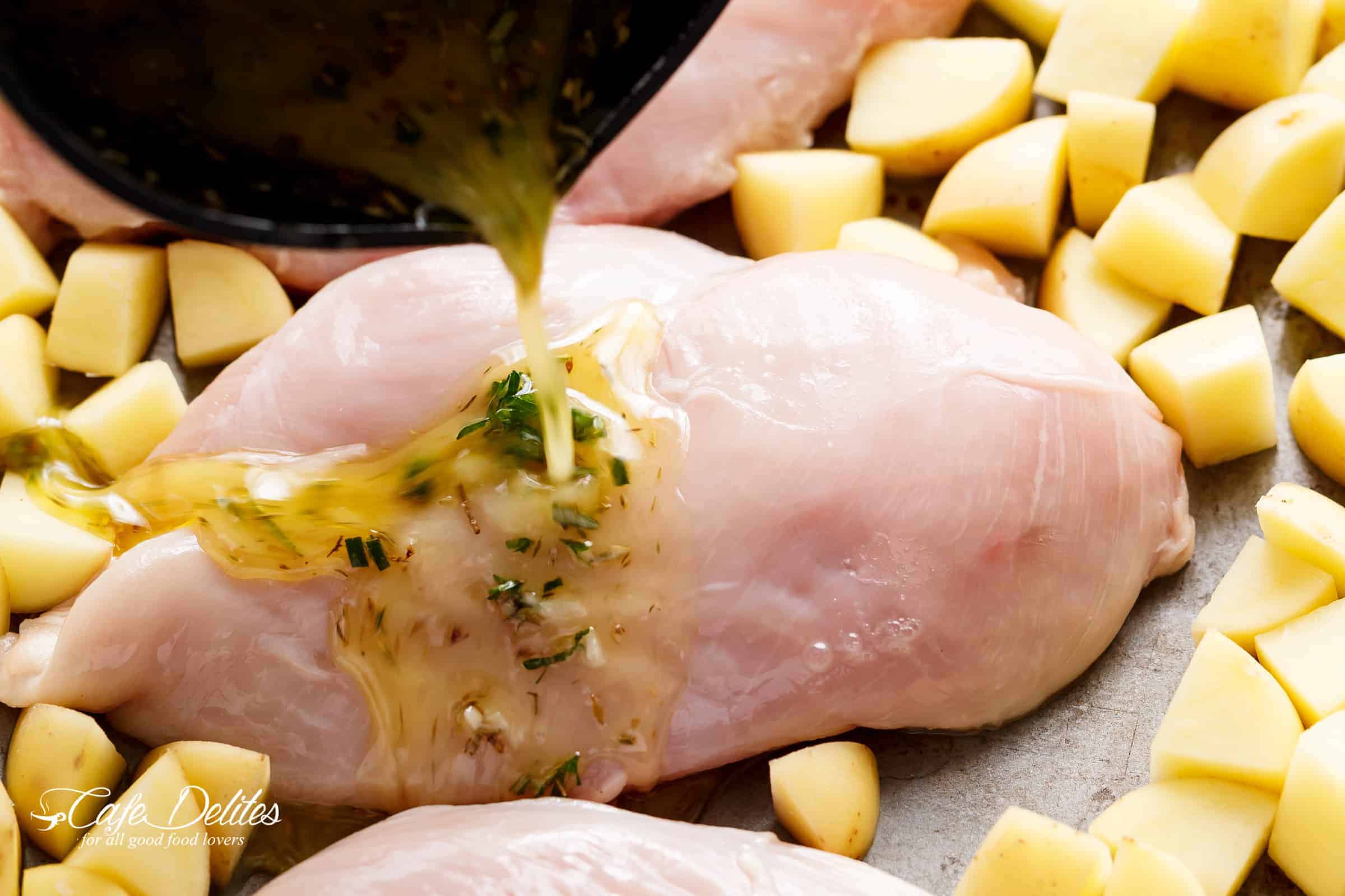 raw chicken breast surrounded by uncooked potatoes on a sheet pan with garlic sauce being poured over top. 