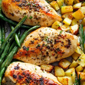 close-up image of baked chicken breasts, potatoes, and green beans. The chicken breasts are arranged in a row, each one glistening with a savory glaze and speckled with herbs. The golden-brown roasted potatoes are scattered around the chicken, adding a hearty and comforting element to the dish. Fresh green beans provide a vibrant pop of color and a hint of freshness.