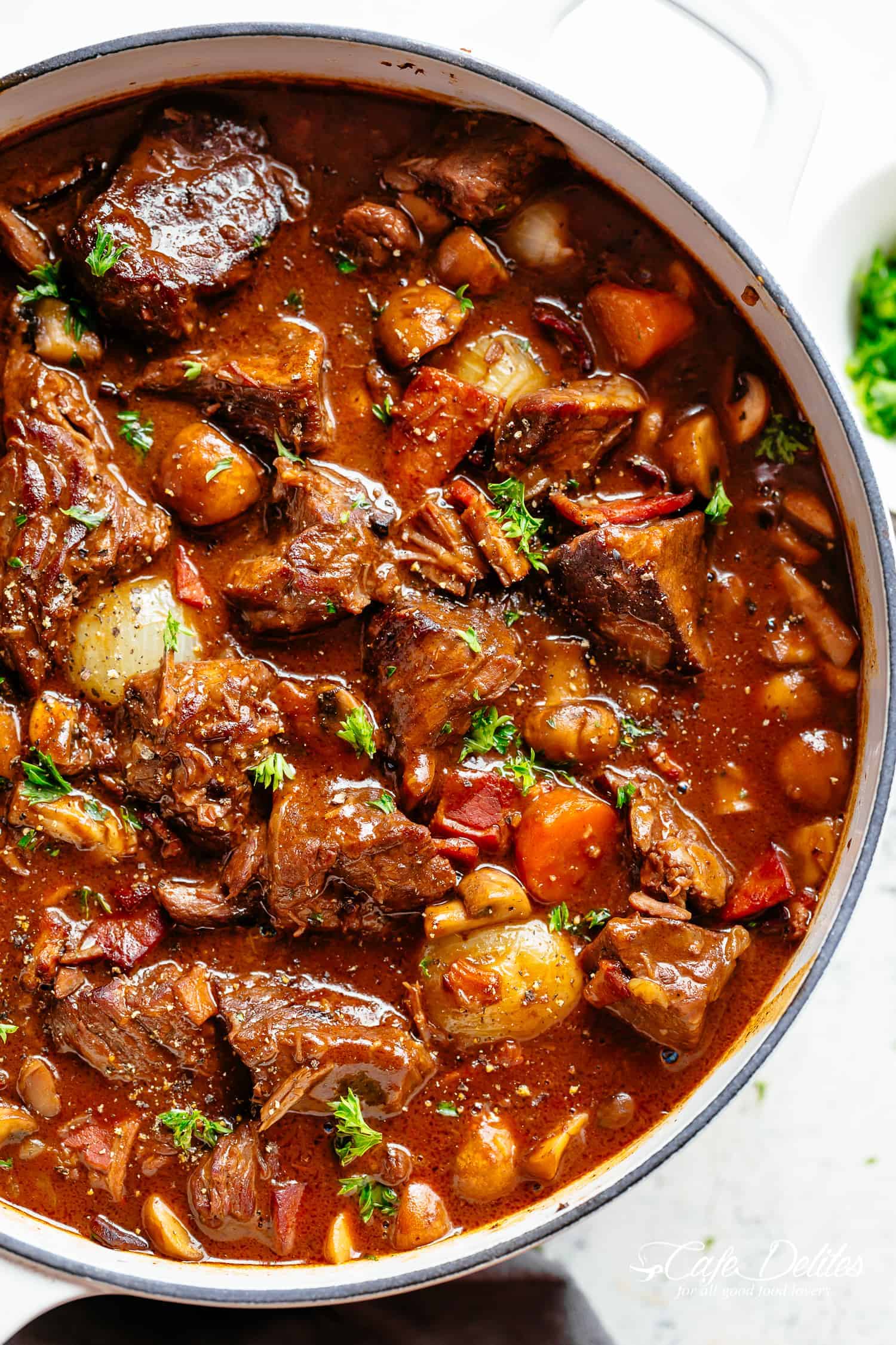 Tender fall apart chunks of beef simmered in a white cast iron casserole dish against a white background. Simmered in a rich red wine gravy, Julia Child's Beef Bourguignon an incredible family dinner. Slow Cooker, Instant Pot/Pressure Cooker, Stove Top and the traditional Oven method included! Easy to make, every step is worth it | cafedelites.com