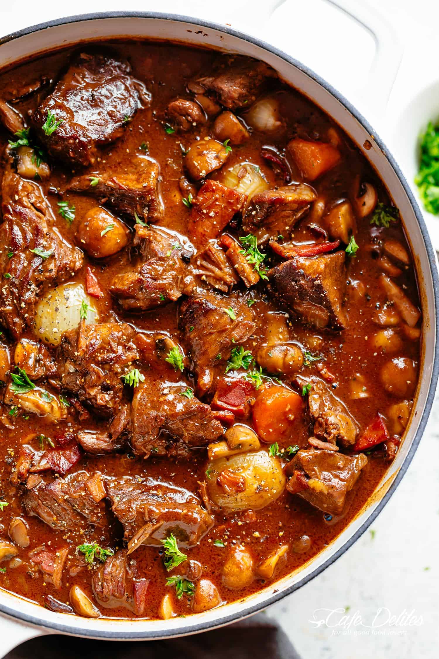 Tender fall apart chunks of beef simmered in a white cast iron casserole dish against a white background. Simmered in a rich red wine gravy.