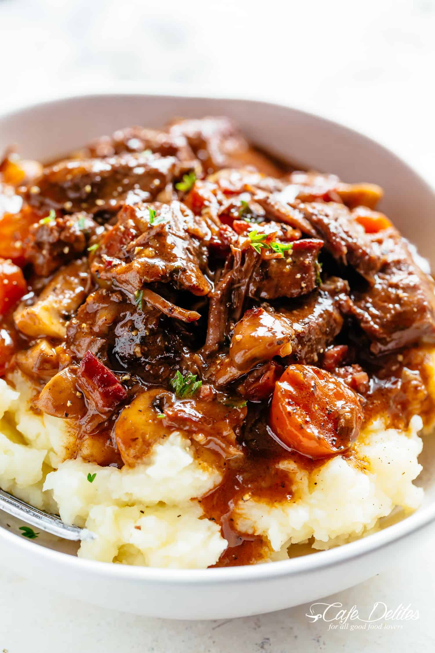 Beef Bourguignon on mashed potatoes with gravy served in a white bowl with a silver fork in a white bowl on a white table