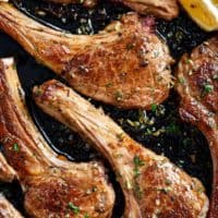 cooked lamb chops in a black pan, green herbs sprinkled on top with two lemon wedges to one side.