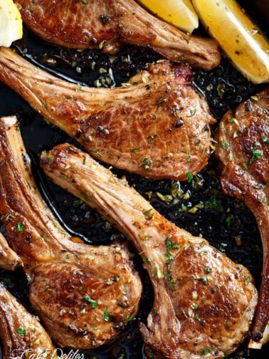 cooked lamb chops pieces in a black pan, green herbs sprinkled on top with two lemon wedges to one side.