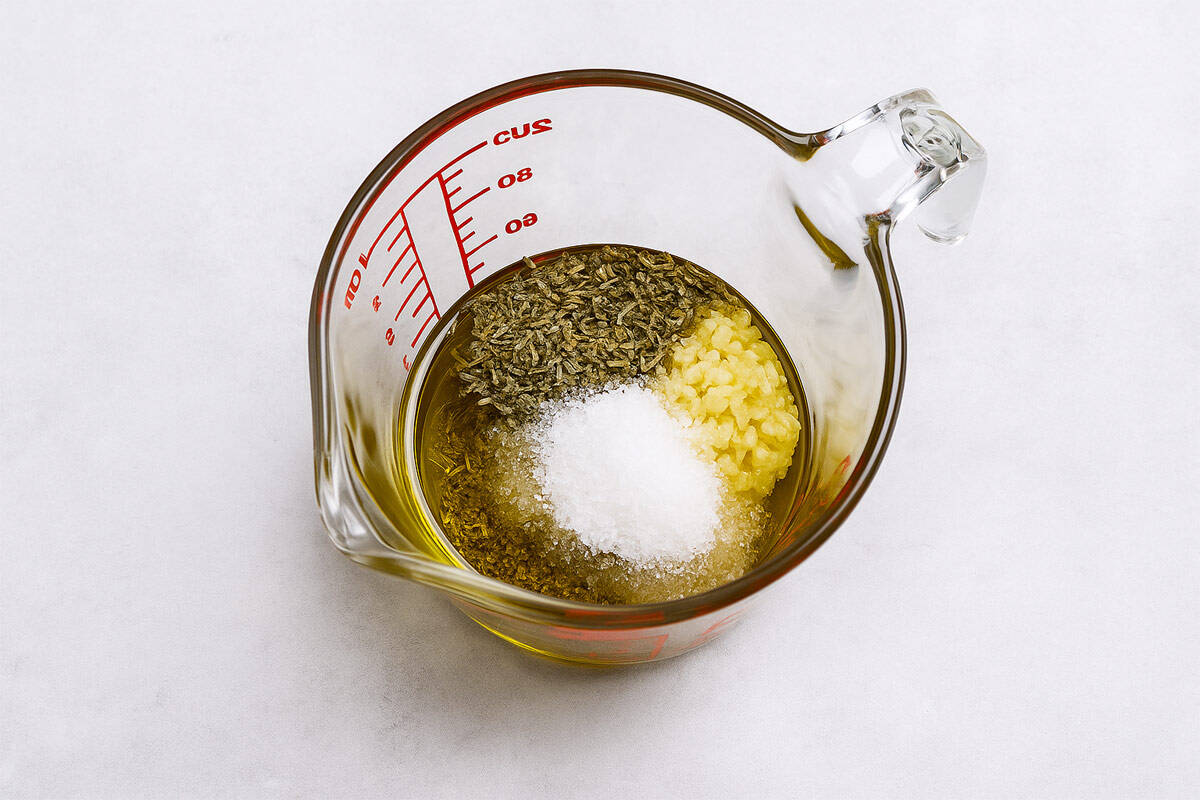 Overhead view of olive oil, oregano, garlic, salt, and pepper layered in a glass measuring cup on a light stone surface.