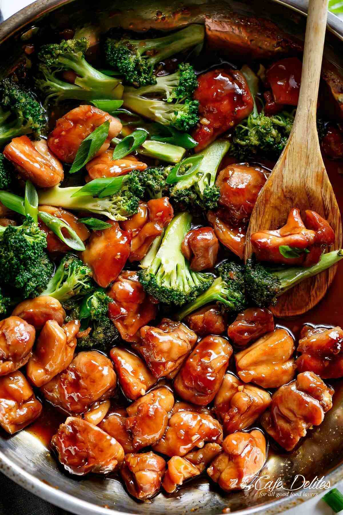 Close-up image of Teriyaki Chicken with broccoli and chives in a pan, being stirred by a wooden spoon soaked in teriyaki sauce.