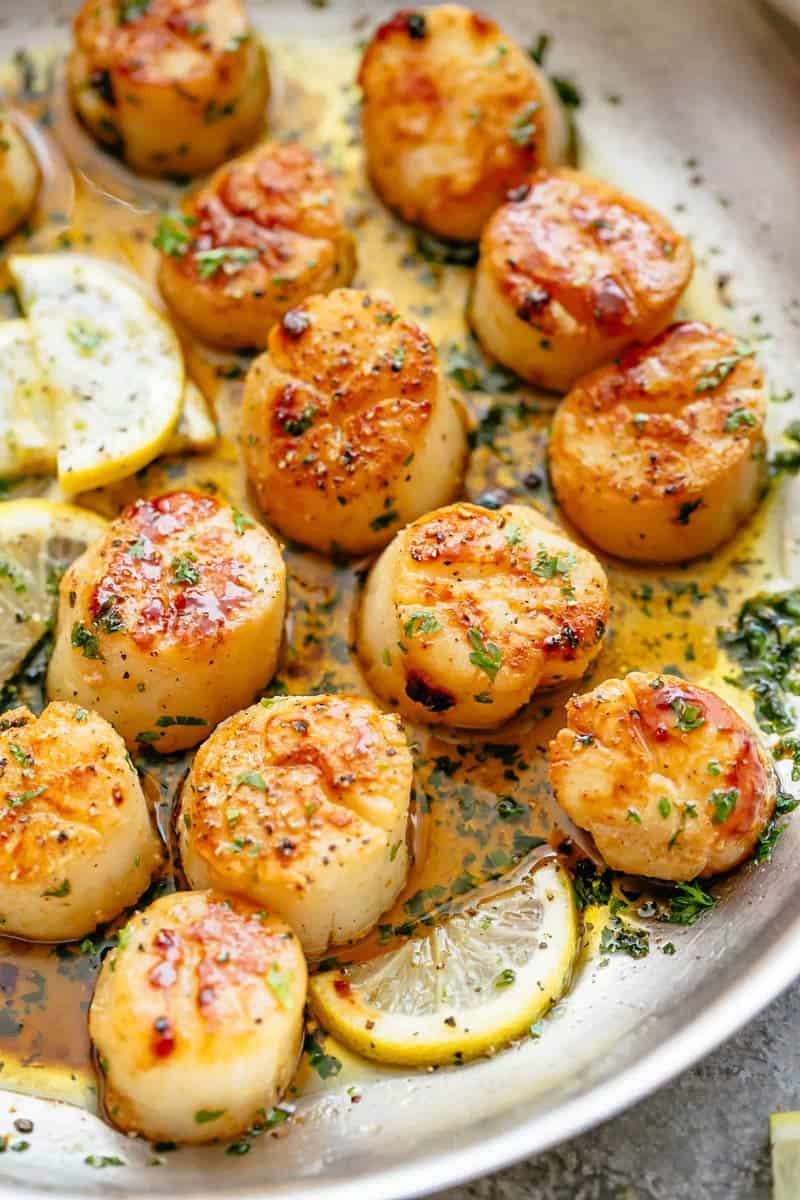 Golden, seared scallops in a pan with a shallow layer of garlic butter, garnished with herbs and several lemon wedges to one side. 