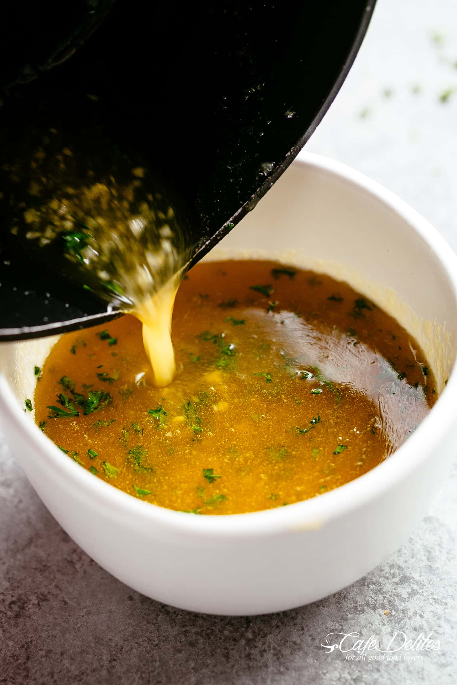 Golden Honey Garlic Butter Sauce pouring from a black pot into small white sauce cup with small bits of garlic and parsley. 