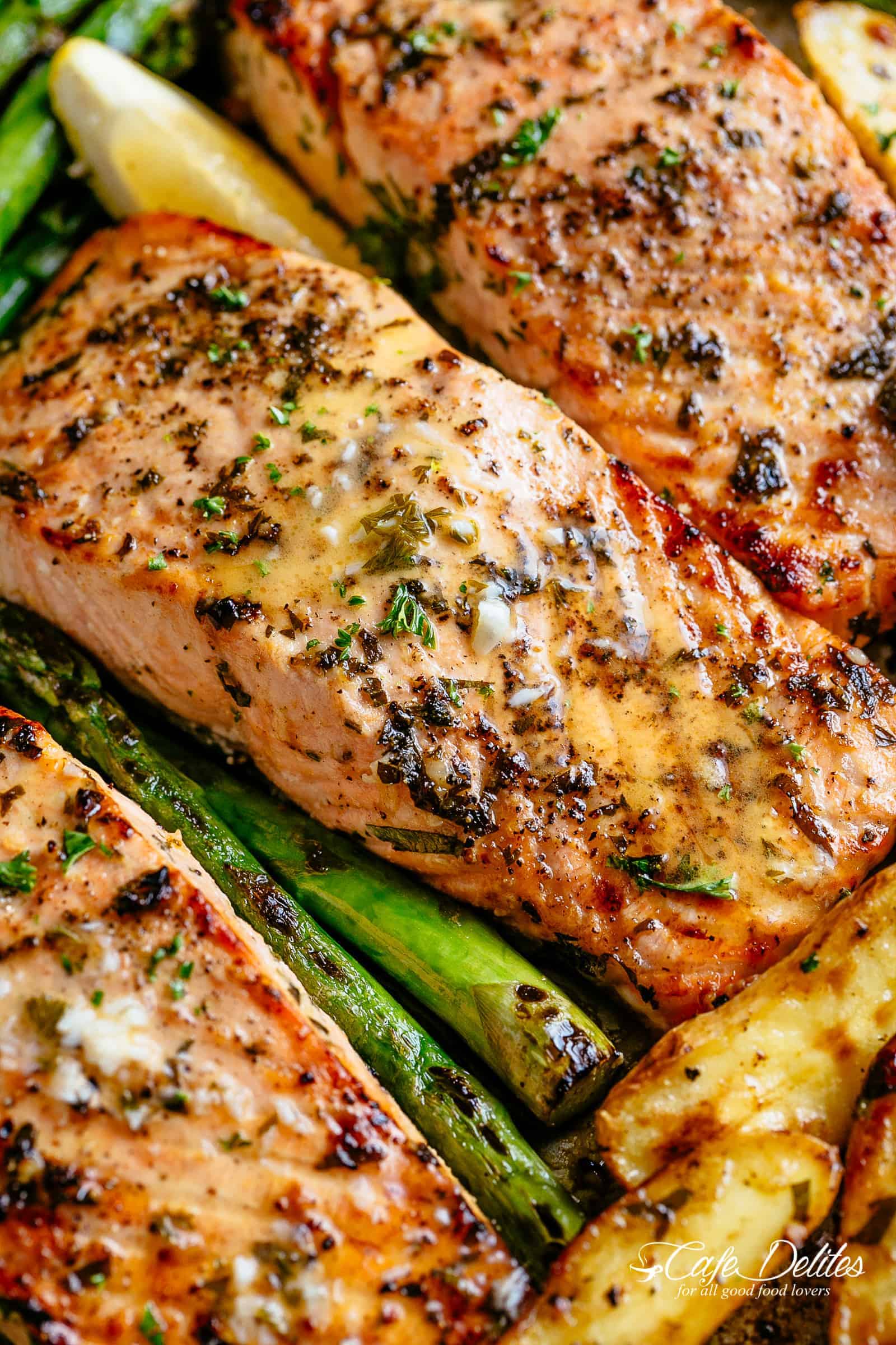 Close up of salmon fillet coated in garlic butter sauce and sprinkled with fresh green herbs. Asparagus and potato wedges are visible from underneath the fish. 