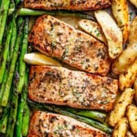 The image shows four salmon fillets on a baking sheet, along with asparagus spears and potato wedges. The salmon is covered in a brown glaze and garnished with fresh herbs and lemon slices. The bright green asparagus is soft and the potatoes are slightly browned, after being baked.