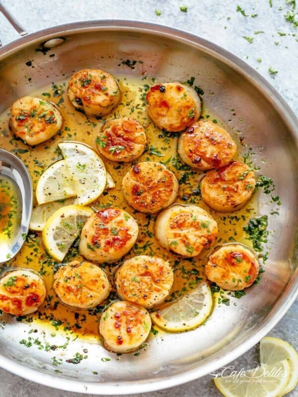 Golden, seared scallops in a pan with shallow layer of garlic butter, garnished with herbs and several lemon wedges to one side.
