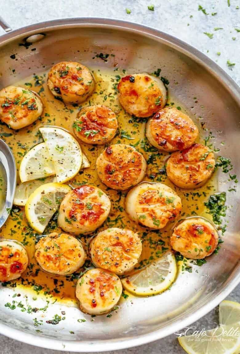 Golden, seared scallops in a pan with shallow layer of garlic butter, garnished with herbs and several lemon wedges to one side.