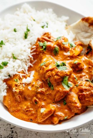 A close-up shot of Butter Chicken, generously garnished with fresh coriander, alongside a bed of fluffy white rice and a piece of naan bread. Chunks of chicken, coated in the flavorful sauce, are scattered throughout the dish.