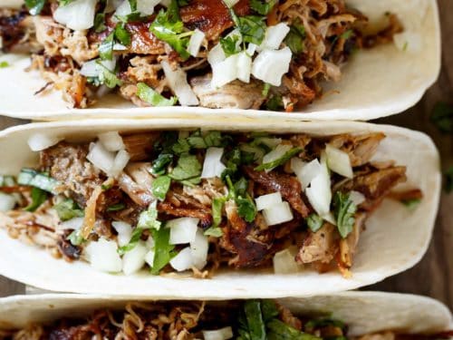 The image shows four corn tortillas, each filled with carnitas, chopped white onion, and chopped cilantro. The tortillas are laid out in a row, slightly overlapping, on a wooden table or cutting board.