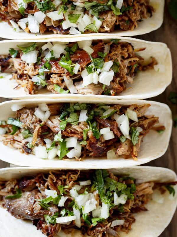 The image shows four corn tortillas, each filled with carnitas, chopped white onion, and chopped cilantro. The tortillas are laid out in a row, slightly overlapping, on a wooden table or cutting board.