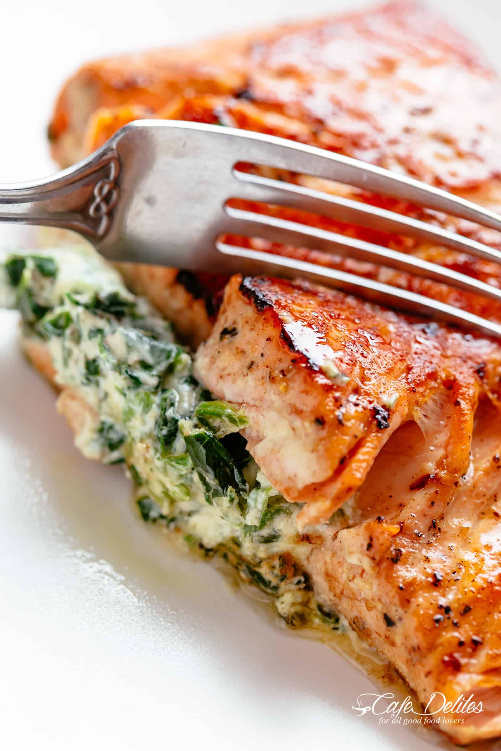 A fork slices through the side of a stuffed salmon fillet on a white plate. Filled with cream cheese, spinach, parmesan cheese and garlic. 