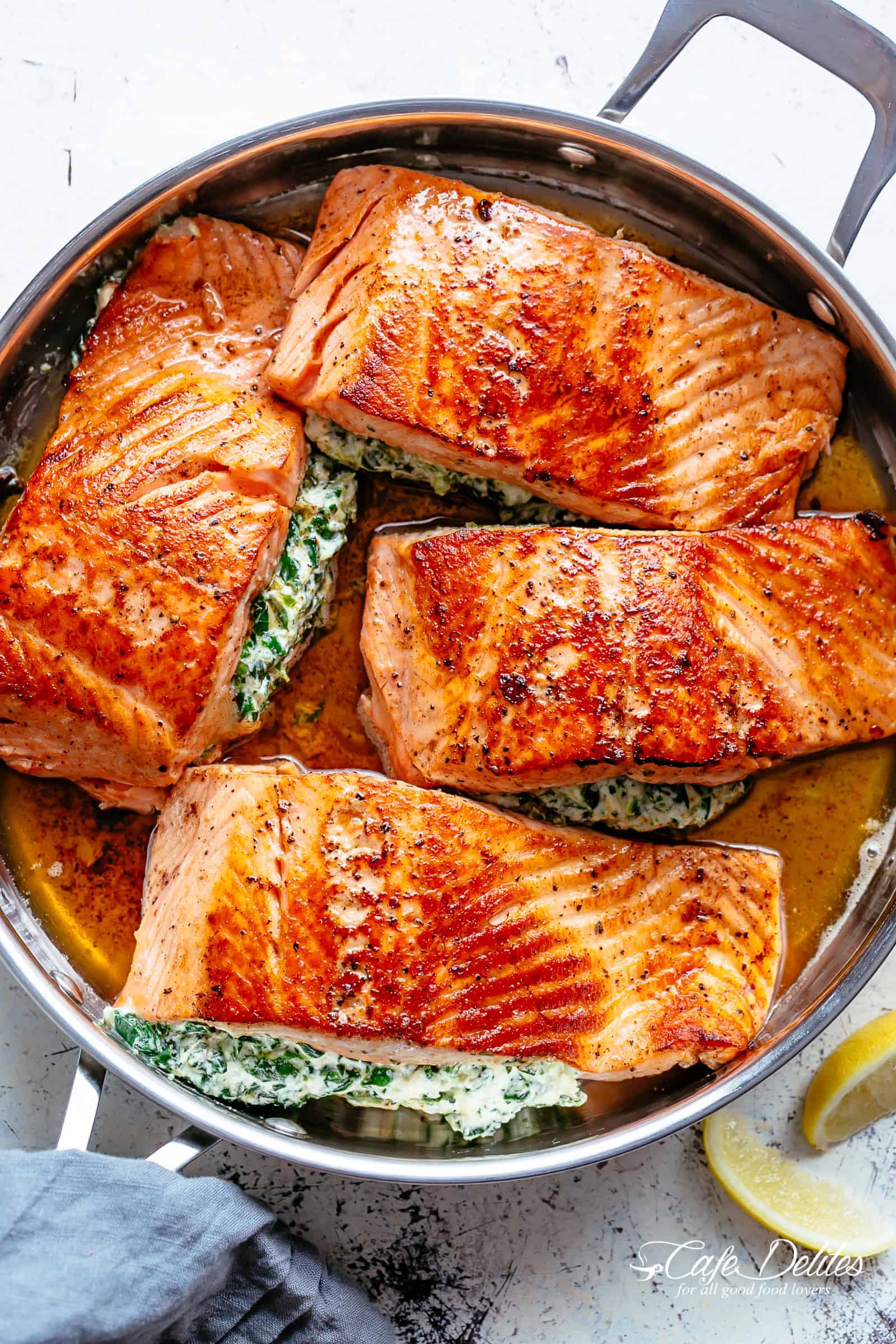 Four Salmon fillets stuffed with creamed spinach in a garlic butter sauce in a silver frying pan served with lemon wedges on a white table.