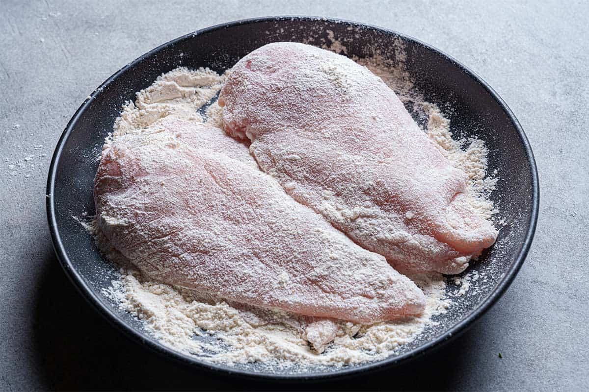 An image of chicken breasts being dipped into a mixture of flour, salt, garlic powder, and pepper in a shallow bowl.