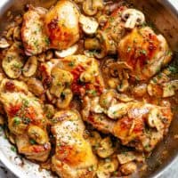 Large steel pan with multiple browned chicken thighs covered in sliced mushrooms and fresh, green herbs.