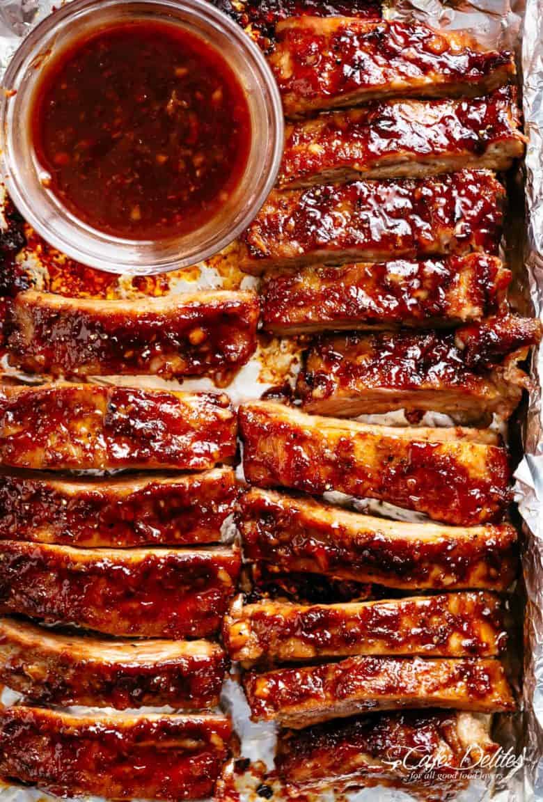 The image shows a top-down view of a baking sheet filled with delicious, glistening ribs cut into individual portions. They are coated in a rich, dark barbecue sauce that has caramelized slightly during cooking, creating a mouthwatering sheen. A small clear bowl sits at the top left corner of the baking sheet, containing more of the sauce, ready to be drizzled over the ribs. The ribs are arranged in an organized manner, slightly overlapping each other, on aluminium foil.