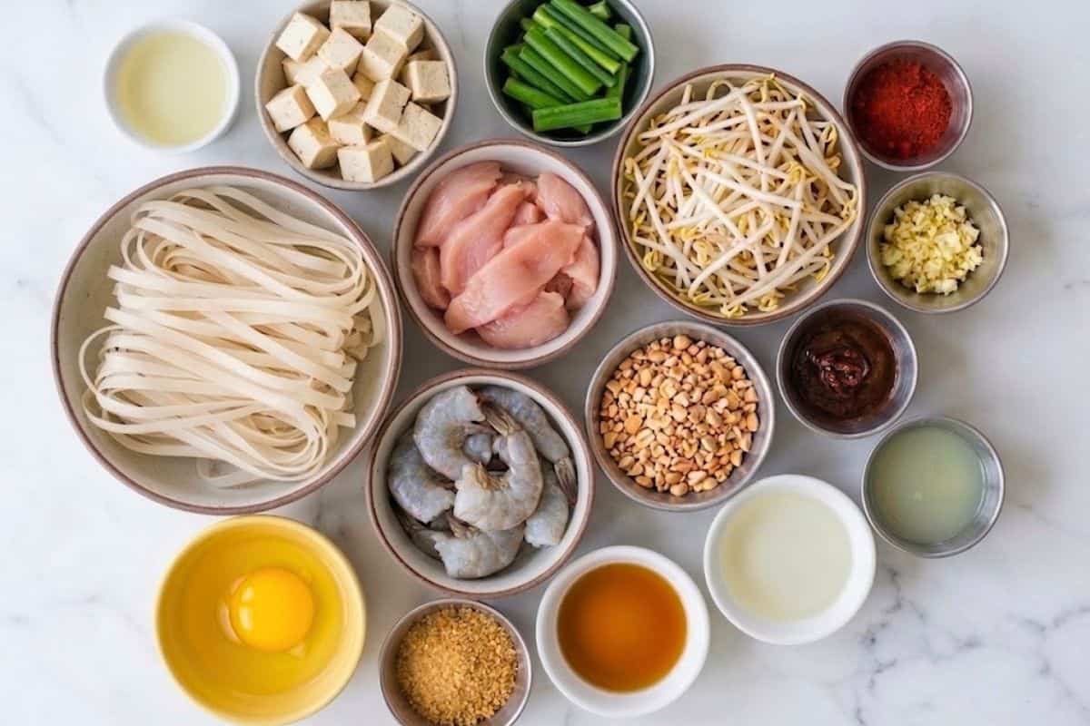 Flat lay photo of ingredient shot for Pad Thai (dried rice noodles, fish sauce, brown sugar, tamarind paste, lime juice, rice vinegar, vegetable oil, minced garlic, diced tofu, egg, boneless skinless chicken breast, large shrimp, paprika, green onion, unsalted chopped peanuts, bean sprouts