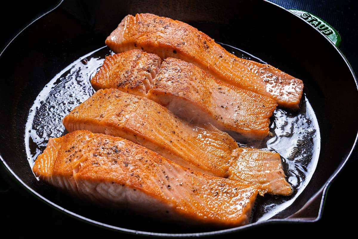 An image of salmon fillets being seared in a pan and seasoned with salt and pepper.