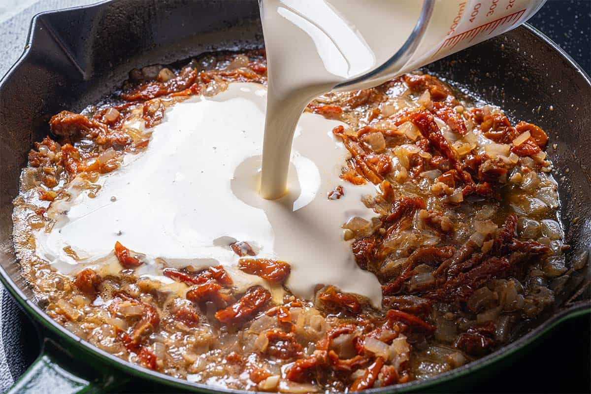 An image of heavy cream being added to the mixture of onions and sun-dried tomatoes.