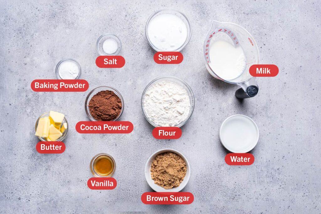 Ingredients laid out against a white background, baking soda, salt, sugar, milk, cocoa powder, flour, vanilla, brown sugar, water