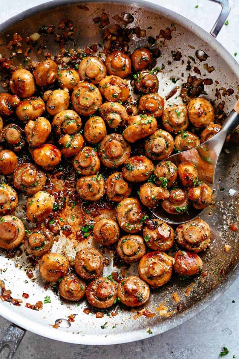 Image of button mushrooms coated in garlic butter sauce, simmering in a pan, ready to be scooped with a kitchen spoon.