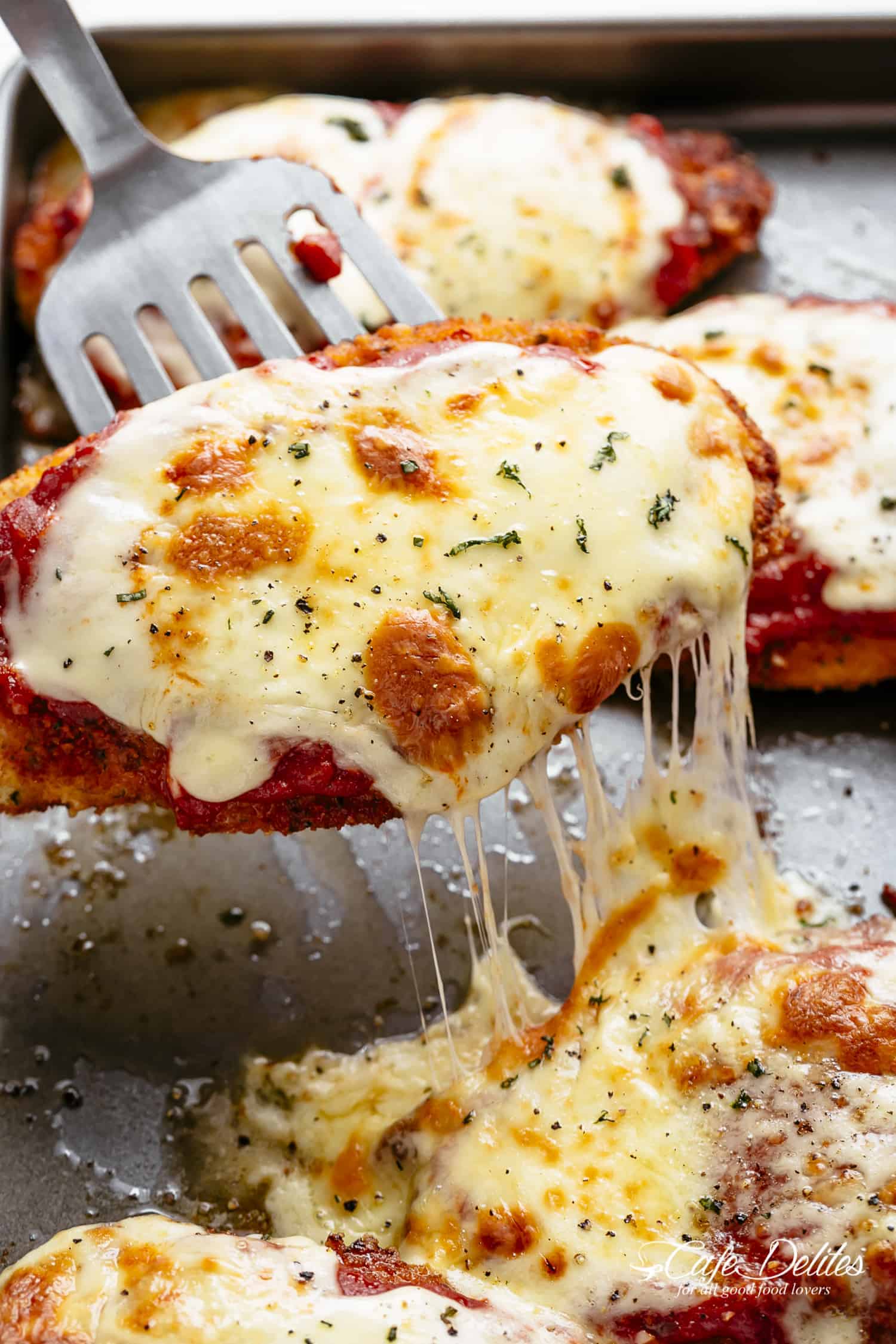 The Best Chicken Parmesan with a deliciously crispy breadcrumb coating, smothered in a rich homemade tomato sauce and melted mozzarella cheese! This is here best Chicken Parmesan you will ever make! One chicken parmesan being lifted from a baking sheet with a metal spatula and lots of stringy, melted cheese. Just fresh out of the oven.