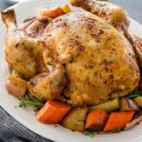 A large slow cooked, golden chicken on a bed of roughly chopped carrots, potatoes and herbs, on a white serving platter.